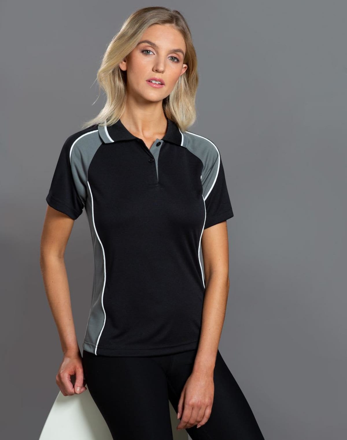 A woman with long blonde hair wearing a black and gray athletic polo shirt and black pants poses against a gray background. She is leaning on a white object, looking directly at the camera.