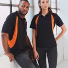Two people in black polo shirts with orange stripes stand indoors near a window. One leans against a wall, while the other stands next to them. Both are wearing casual jeans.