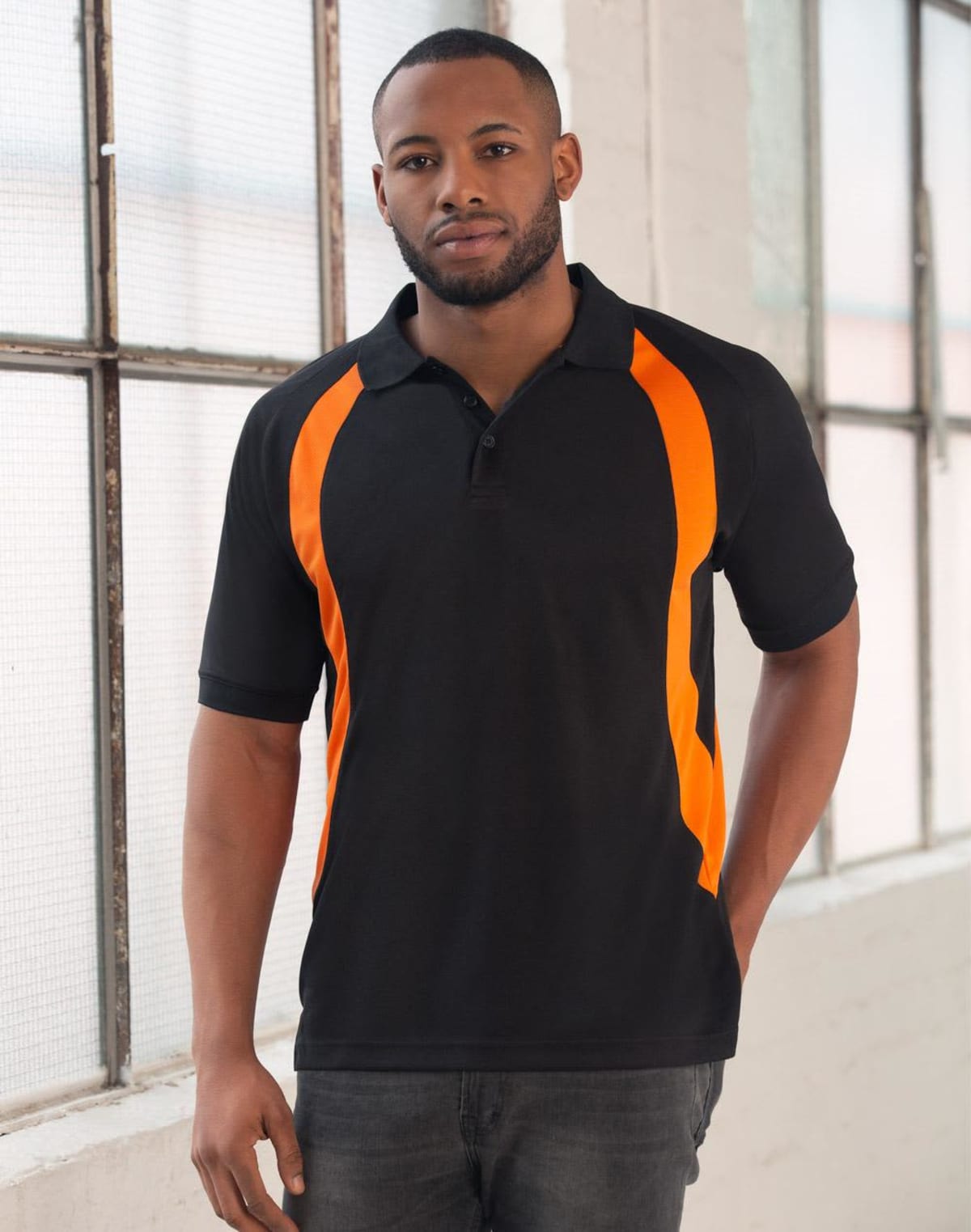 A man standing indoors wearing a black and orange polo shirt. He is in front of a large industrial-style window with soft light coming through. The man has a neutral expression and is casually resting one hand in his pocket.