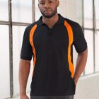 Person wearing a black polo shirt with orange stripes stands indoors near large windows, posing with one hand in pocket. Casual attire with a relaxed expression.