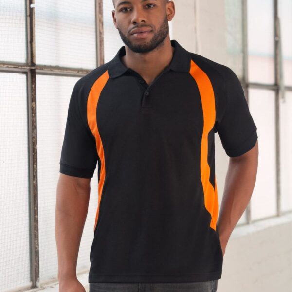 Person wearing a black polo shirt with orange stripes stands indoors near large windows, posing with one hand in pocket. Casual attire with a relaxed expression.