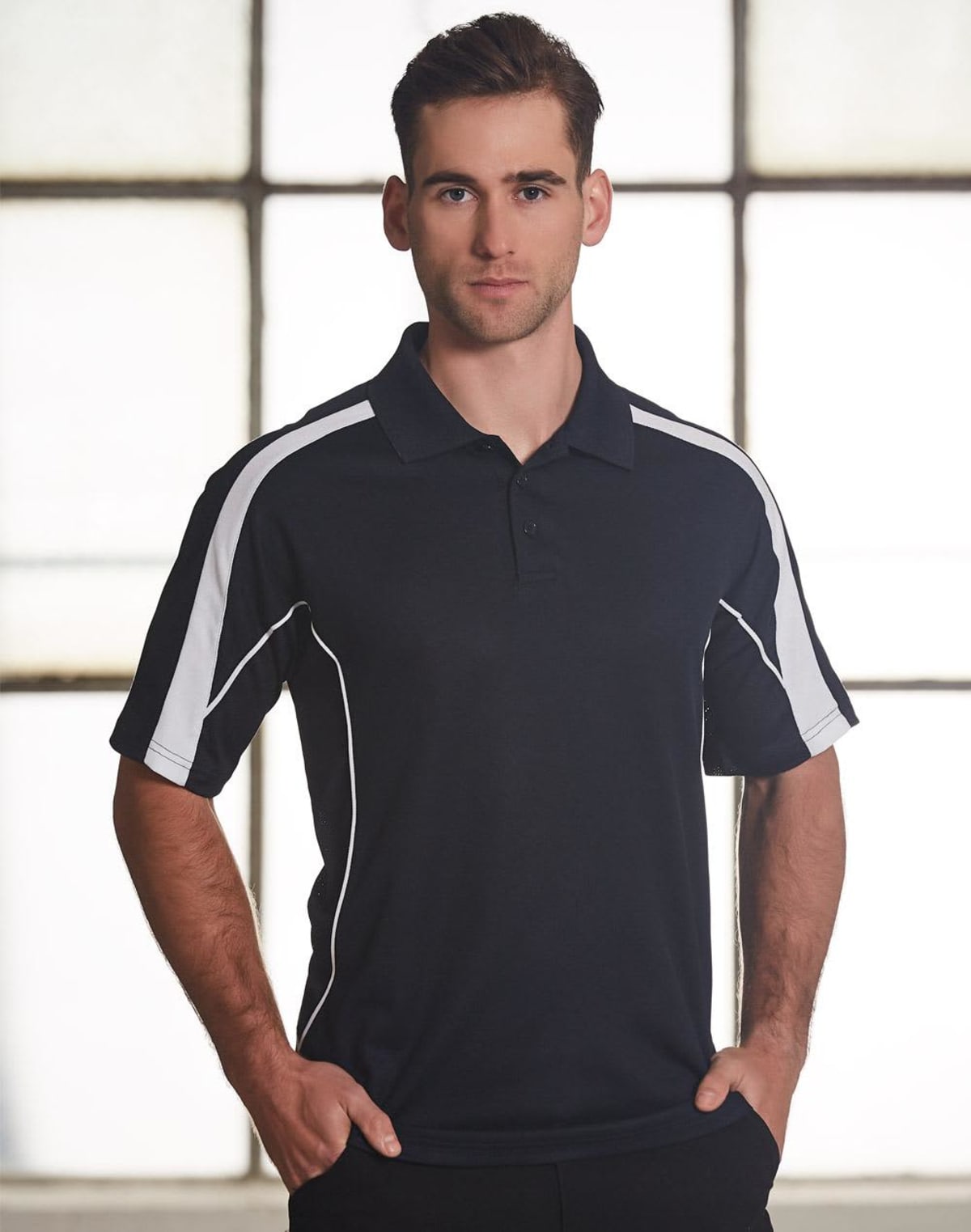 A man standing in front of a large window wearing a black polo shirt with white stripes on the shoulders. He has short brown hair and is looking directly at the camera with a neutral expression.
