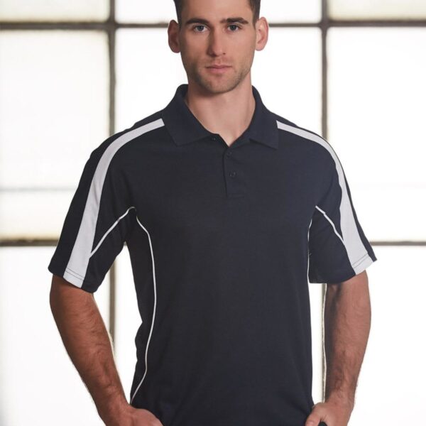 A man stands in front of a large window wearing a black polo shirt with white stripes on the sleeves. His hands are in his pockets, and he has short dark hair. The background is softly lit.