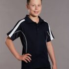 A young boy with short hair is smiling and standing with one hand on his hip. He is wearing a black polo shirt with white stripes on the shoulders. The background is plain gray.