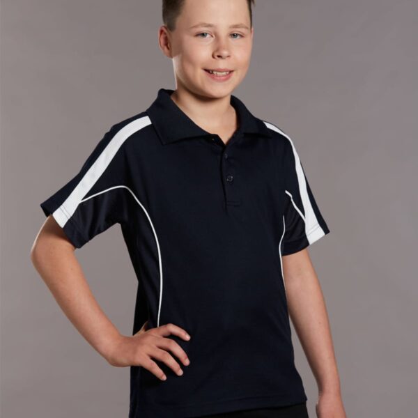 A young boy with short hair is smiling and standing with one hand on his hip. He is wearing a black polo shirt with white stripes on the shoulders. The background is plain gray.