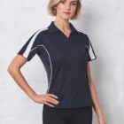 A person stands against a white wall wearing a black and white short-sleeved polo shirt and black pants. They have short, wavy blonde hair and are posing with one hand on their hip, looking directly at the camera.