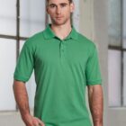 A person wearing a green polo shirt and black pants stands indoors against a blurred background of large windows and a white wall.