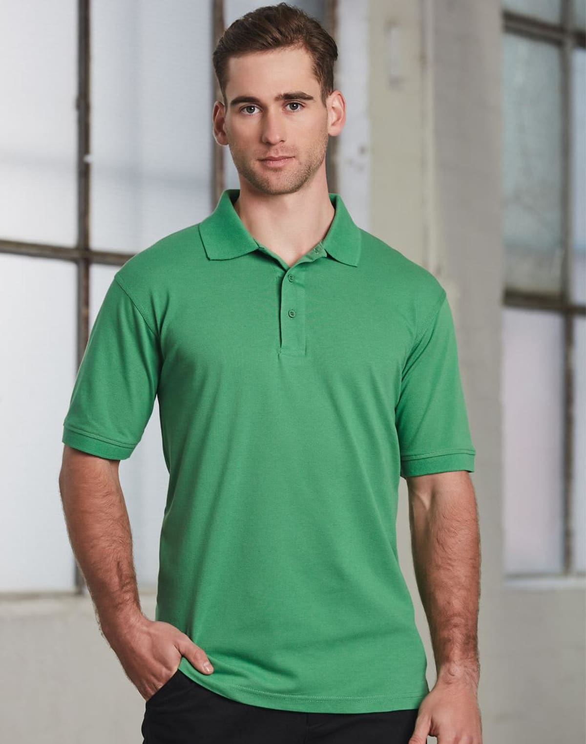 A person wearing a green polo shirt and black pants stands indoors against a blurred background of large windows and a white wall.