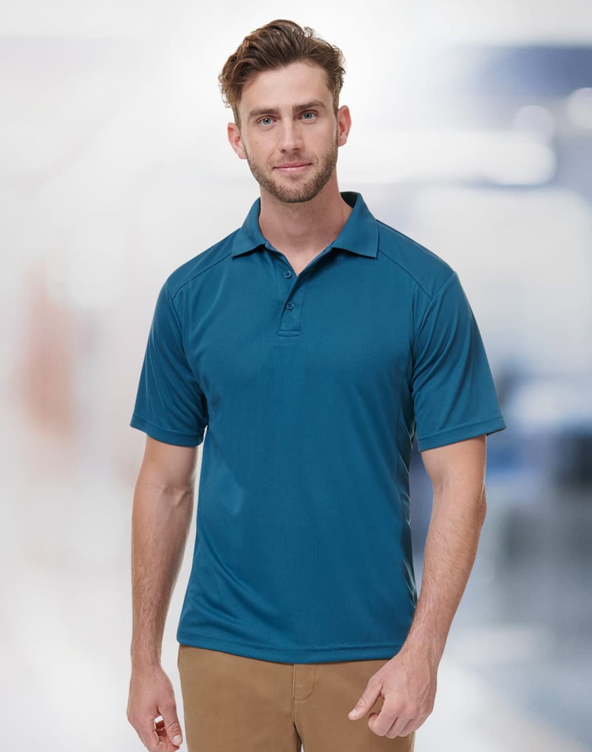 A man with short hair, wearing a teal polo shirt and beige pants, standing in a well-lit space with a blurred background.