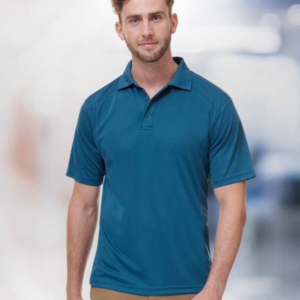 A man with short hair wears a teal polo shirt and tan pants. He stands against a blurred background, facing the camera with a neutral expression.