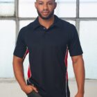 A person standing indoors, wearing a black polo shirt with red and white side panels. The background features a large window with a textured glass design. The person has their hands in their pockets and is looking at the camera.