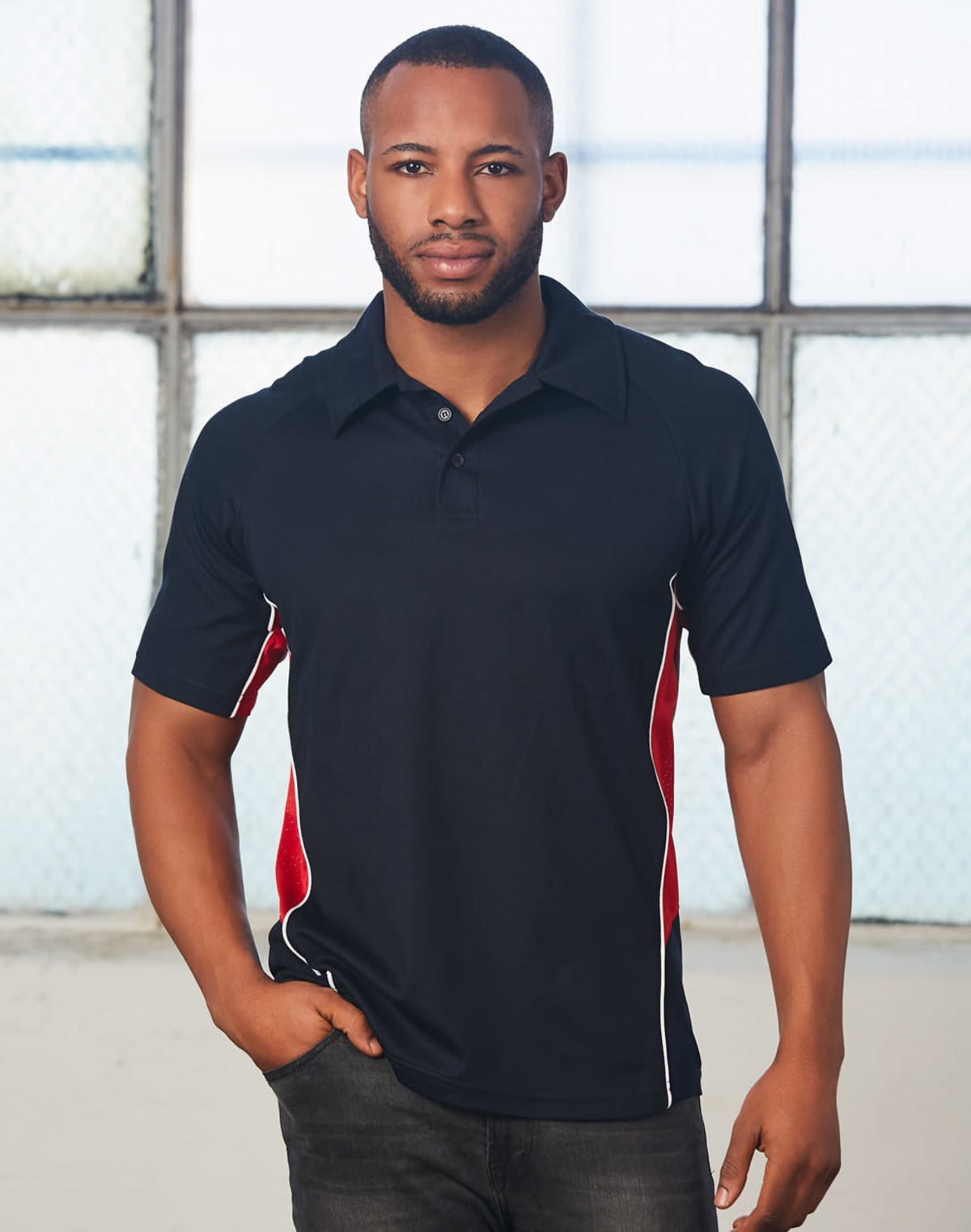 A person standing indoors, wearing a black polo shirt with red and white side panels. The background features a large window with a textured glass design. The person has their hands in their pockets and is looking at the camera.