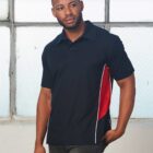 A man stands in front of a large, grid-patterned window. He is wearing a black polo shirt with red and white side panels and dark jeans. Hes looking slightly to the side with a neutral expression.