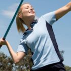 Person in a light blue polo swinging a long stick outdoors under a clear blue sky.