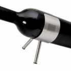 A wine bottle placed horizontally on a minimalistic, silver metal stand with two legs. The bottle appears dark with a cork closure and a modern design. The background is plain white.