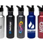 Five stainless steel water bottles with black lids and straws, each in a different color: silver, black, dark gray with a multicolored logo, blue with a white flame logo, and red with MQ logo. Each bottle has a unique company logo printed on it.
