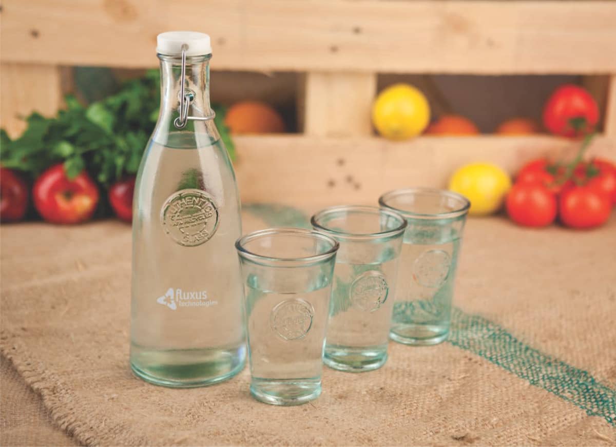 A clear glass bottle filled with water is placed next to three empty glasses on a burlap cloth. In the background, wooden crates contain a mix of fresh tomatoes and other produce.