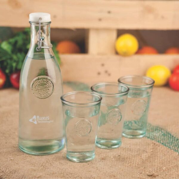 A glass bottle filled with water is placed on a burlap surface next to three empty glasses. In the background, there are assorted fruits and vegetables, including tomatoes and peppers, displayed on a wooden crate.