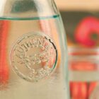 Close-up of a glass bottle with an embossed logo reading Authentic Glass in a circular design. The background is slightly blurred, showing a red bell pepper and a couple of glasses. The lighting is warm and inviting.