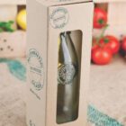 A glass bottle in eco-friendly cardboard packaging with a cut-out window. The box is labeled Authentic 100% Recycled Glass. Fresh tomatoes and a wooden crate are in the blurred background.