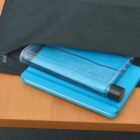 A black bag is partially open on a wooden table, revealing a flat, rectangular water bottle and a blue notebook inside.