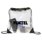 Transparent drawstring bag with FOXTEL logo. Visible inside are a smartphone, wallet, lotion tube, keys, and a small purple item. The bag has black corners and strings.