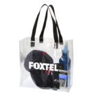 Clear tote bag with black handles, containing a dark cap, a sunscreen bottle, a tube of cream, and a small electronic device. The bag has FOXTEL printed on the front in bold white letters.