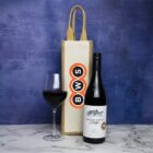 A bottle of Keenans Bridge Shiraz stands next to a full wine glass and a BWS-branded wine bag. They are displayed on a marble surface against a textured blue background.