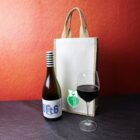 A bottle of wine with a striped cap labeled the 6 is just the limit stands next to a filled wine glass. Behind them is a beige canvas bag with a green emblem on a dark slate surface and red background.