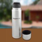 A silver thermos with a black lid, featuring the amazon.com logo, stands upright on a brown surface. The cap is removed and placed next to the thermos. A blurred outdoor background is visible.