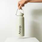 A hand holding a light green water bottle with a handle against a white background. The bottle features the word Aēsop in black lettering. It is held above a plain white surface.