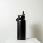 Black insulated water bottle with a handle and flip-top lid, placed on a white surface against a plain light-colored background.