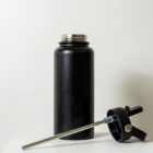 A black insulated water bottle with its lid removed sits on a white surface. The lid, featuring a straw, is placed next to the bottle. The background is a plain white wall.
