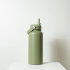 A green water bottle with a lid featuring a flip-up spout sits on a white surface. The bottle has a minimalist design and the background is a plain, light-colored wall.