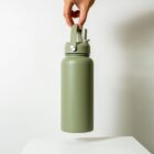 A hand holding a large, olive-green insulated water bottle with a looped handle and a screw-on lid, positioned above a plain white surface. The background is a neutral light gray.