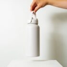 A hand holding a white insulated water bottle by its handle against a plain white background. The bottle is large and cylindrical, with a secure lid.