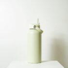 A light green water bottle with a flip-top straw lid sits on a white surface against a plain white background. The bottle has a sleek and simple design.