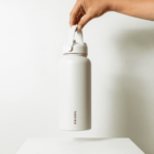 A hand holds a white Prada water bottle with a handle against a plain white background. The bottle is sleek and cylindrical, with the Prada logo vertically printed near the bottom.