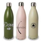 Three insulated water bottles are standing side by side. The first is green with Dove branding, the second is pink with Chando branding and a star graphic, and the third is beige with a Converse All Star logo. Each has a metal cap.