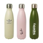 Three insulated stainless steel water bottles with metallic caps. The bottles are cream with a logo, pink with script text, and olive green with bold text, respectively, standing side by side against a white background.