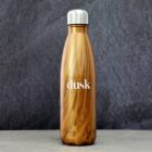 A stainless steel water bottle with a wood grain finish stands upright on a gray stone surface. The word dusk is printed in white on its side. The cap is silver, contrasting with the bottles rich wooden appearance.