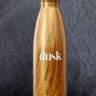 A wooden-textured stainless steel water bottle with a silver cap, standing against a dark gray background. The word dusk is printed in white on the bottle.