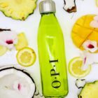 A green glass bottle with a silver cap is surrounded by tropical fruits: slices of pineapple, lemon, mango, and coconut, along with white and pink flowers. The bottle has O.P.I written on it.