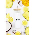 A white tumbler with a wooden lid and metal straw is surrounded by slices of lemon, pineapple, mango, and pieces of coconut arranged creatively on a white background. The tumbler features the Cancer Council logo.