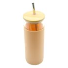 A tall, cylindrical glass tumbler covered with a light peach silicone sleeve. It has a bamboo lid and a stainless steel straw. The tumbler is filled with an orange liquid.