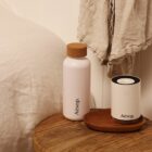 A bedside setup featuring a white Aesop bottle with a cork lid and a small Aesop diffuser on a wooden table. A beige cloth hangs nearby, and part of a bed with white linens is visible on the left.