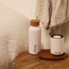 A white Aesop bottle with a cork lid and a matching Aesop cylindrical container sit on a wooden table. A beige cloth hangs nearby. The background features a white wall and part of a bed with a white duvet.