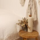 A cozy bedroom setting with a small wooden stool beside a bed. The stool holds a pale beige DIOR bottle and a metal pitcher filled with dried flowers. Neutral tones dominate the decor, with linen bedding and hanging fabric visible.