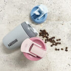 Reusable Nestlé cup with two additional colorful lids (blue and pink) next to scattered coffee beans on a speckled surface.