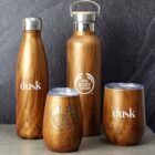 Four wooden textured drinkware items are arranged on a stone surface. From left to right: a dusk bottle, a tall The Body Shop bottle, a The Body Shop tumbler, and a dusk tumbler. Each has a sleek, modern design with brand logos.